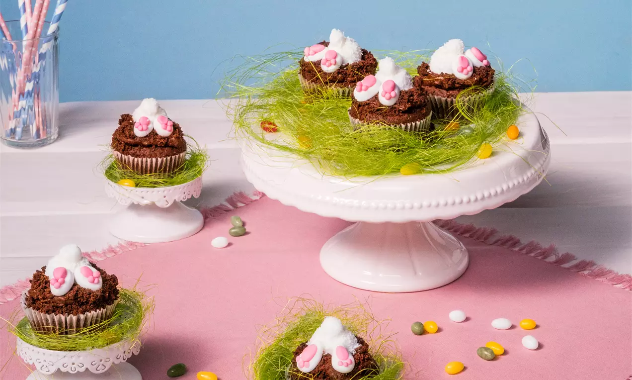 Schoko-Cupcakes mit brauner Krümeldecke, verziert mit weißen und pinken Zucker-Osterhasenfüßchen, auf grüner Zuckerdekokrässe drapiert
