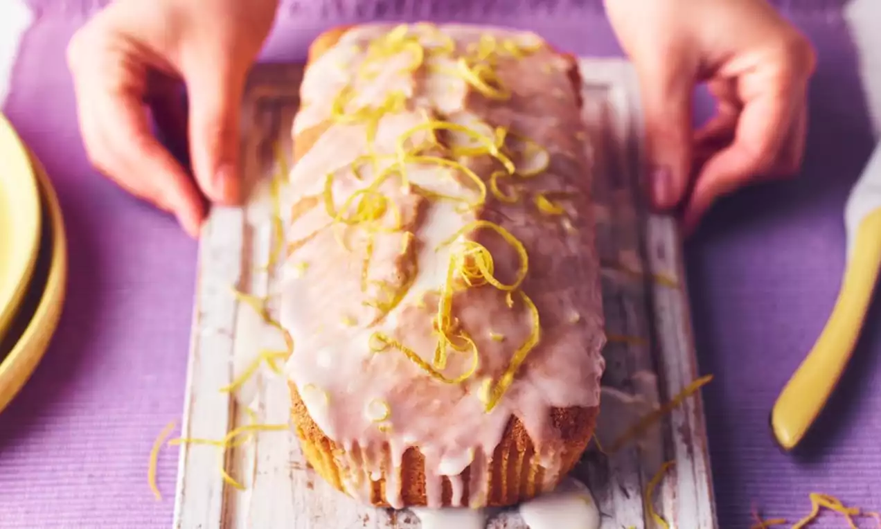 Lemon Madeira loaf cake with golden-brown crust, glossy white glaze dripping down the sides, topped with vibrant yellow lemon zest strips