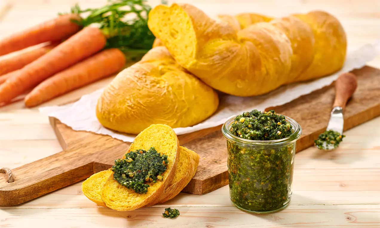 Pesto verde intens într-un borcan alături de felii aurii de pâine moale, pe un platou de lemn, decorat cu morcovi portocalii și frunze