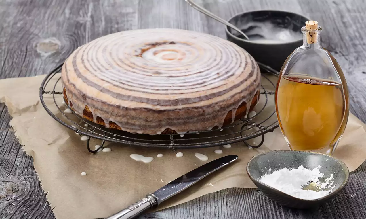 Zebrakuchen mit weißem Guss, kreisförmigem Muster und dunkler Kruste auf einem Gitter, umgeben von Puderzucker, Glasur und Likörflasche