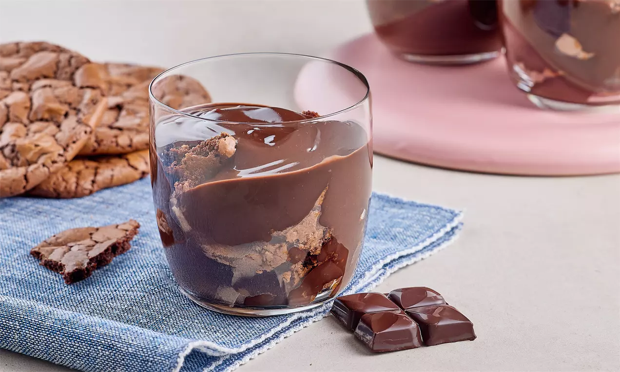 Cam kup içinde yoğun kıvamlı çikolatalı puding ve iri parça çikolatalı kurabiyeler katmanlı şekilde düzenlenmiş, yanında çikolata küpleri
