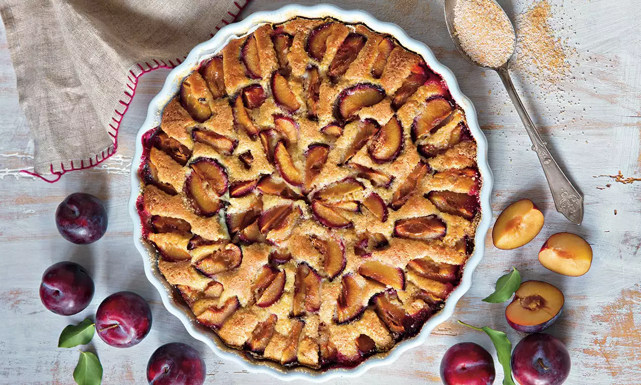 Koláč so zlatistou kôrkou, na povrchu ozdobený vejármi tmavofialových sliviek, v bielej keramickej forme, vedľa čerstvých sliviek a lyžičky s trstinovým cukrom