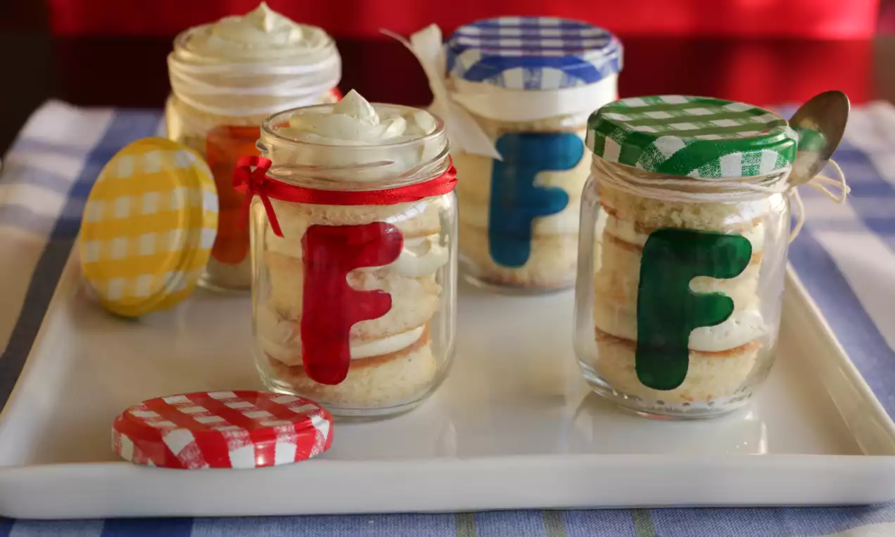 Potinhos de vidro transparentes com camadas de creme claro e biscoito, decorados com fitas e tampas coloridas sobre bandeja branca
