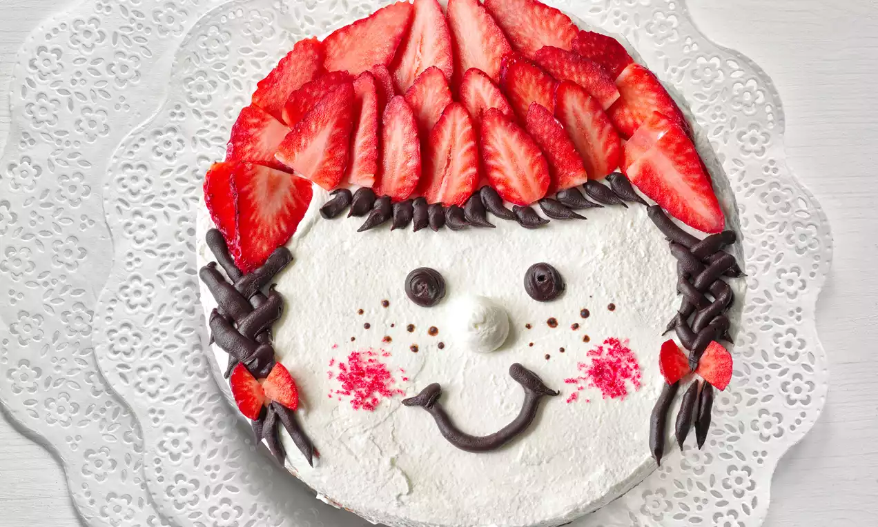 Okrúhla torta s bielym krémovým povrchom, jahodami tvarovanými do vlasov, čokoládovými copmi, červenými líčkami a úsmevom na čipkovanom podklade