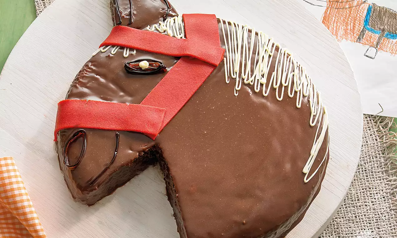Ponykopf-Kuchen mit glänzend brauner Glasur, rotem Zuckergurt, weißer Mähnenverzierung und ovalem Auge auf hellem Holzbrett