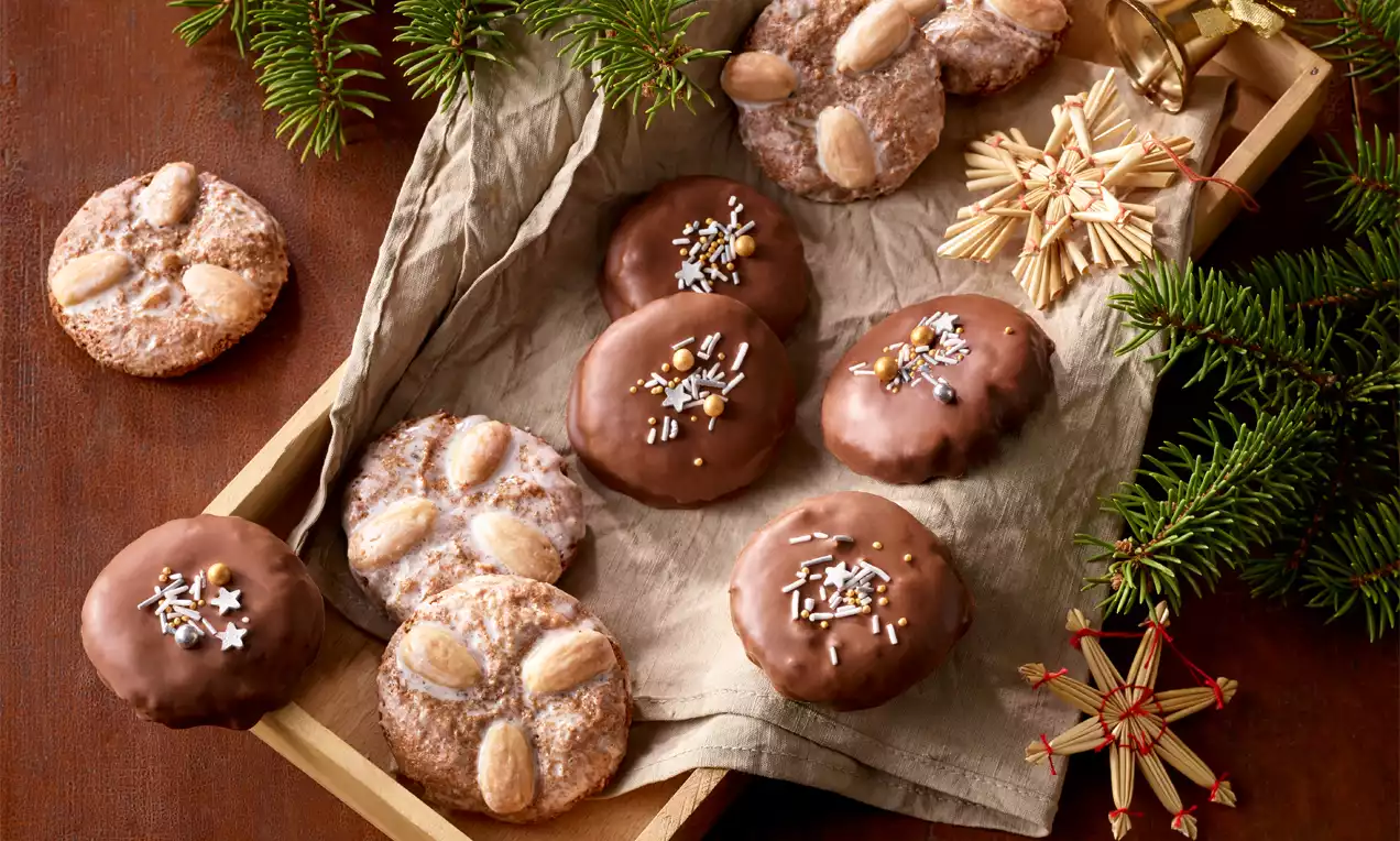 Runde Elisenlebkuchen, teils schokoliert, teils glasiert, mit Mandeln und Streuseln dekoriert, auf Packpapier in einer Holzkiste mit Tannenzweigen.