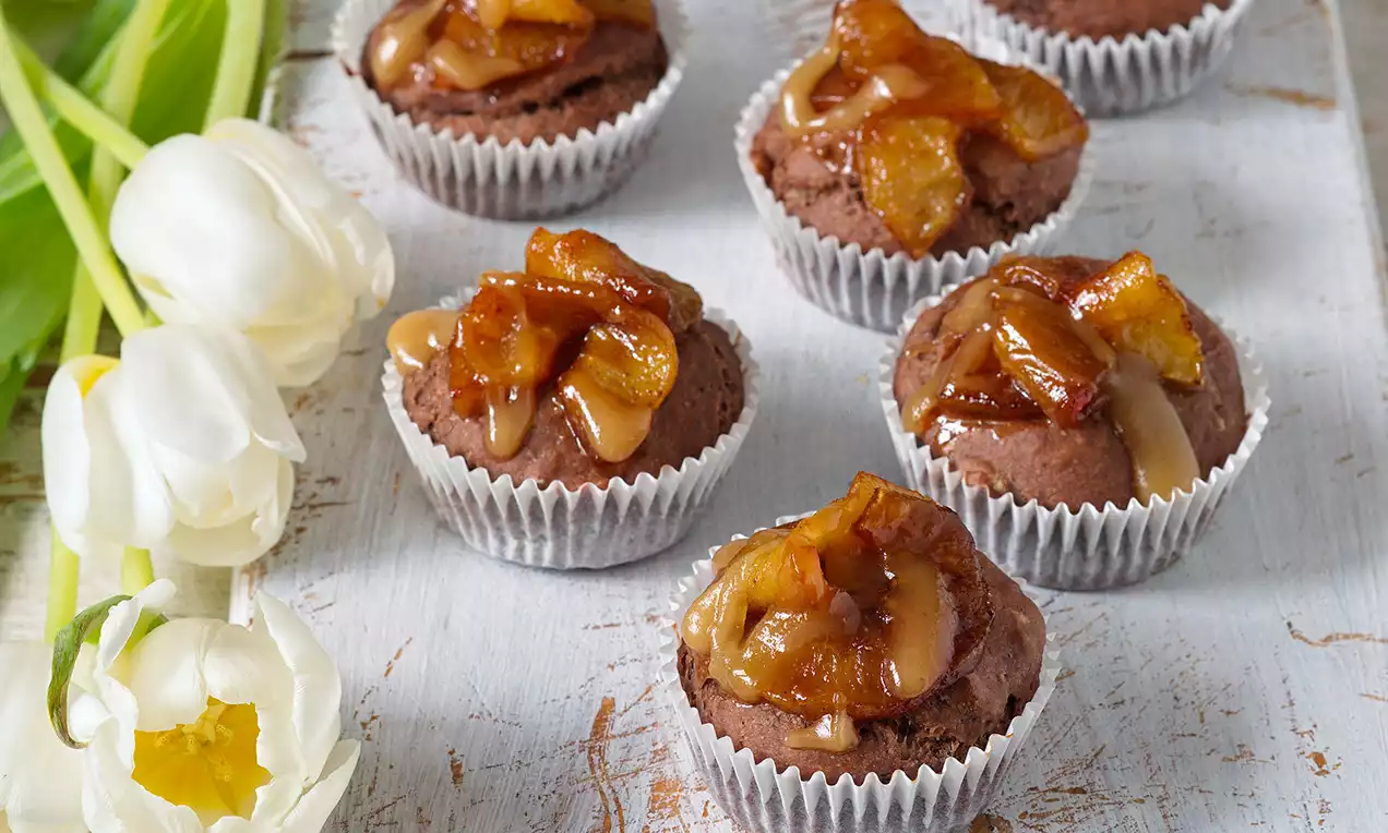 Čokoládové muffiny v bílých papírových košíčcích, ozdobené lesklými glazovanými kousky jablek, na světlém dřevěném podkladu vedle bílých tulipánů