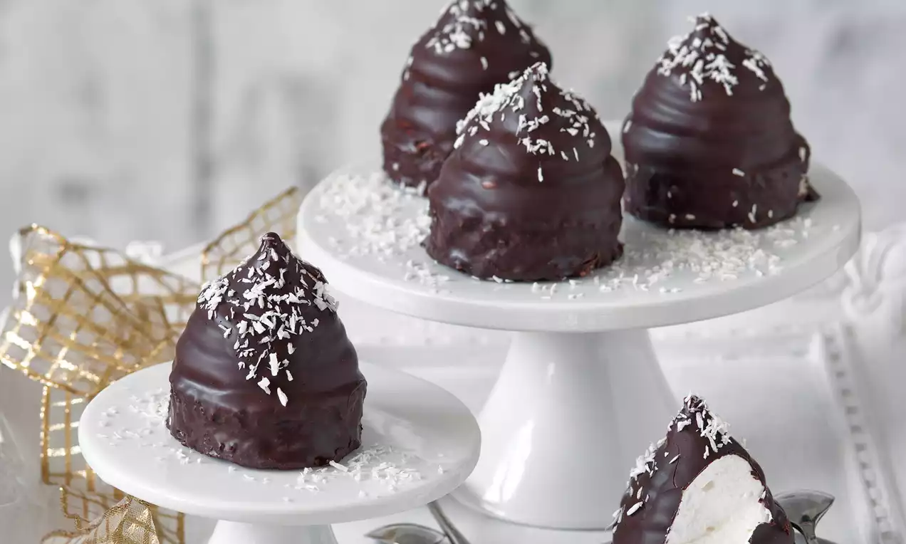 Čokoládové zákusky v tvare kužeľov poliate lesklou tmavou polevou, posypané strúhaným kokosom na bielych podnosoch s jemnou dekoráciou