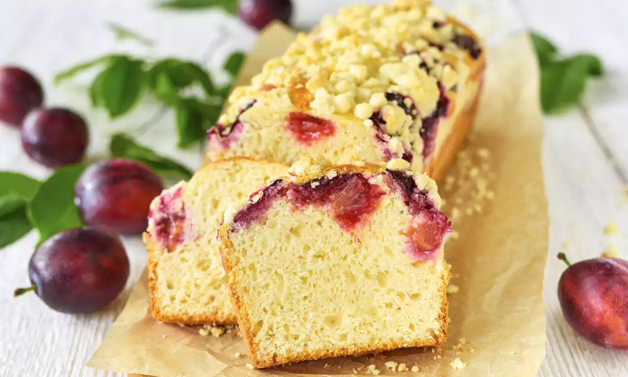 Podłużna babka drożdżowa o złocistej skórce, z soczystymi śliwkami i kruszonką, na pergaminie, otoczona świeżymi śliwkami i zielonymi liśćmi