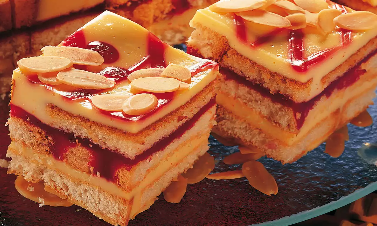 Zweischichtige Kuchenstücke mit hellem Biskuit, roter Fruchtfüllung und gelber Creme, garniert mit gehobelten Mandeln auf Glasplatte