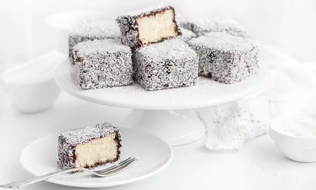 Lamington sponge cakes coated in chocolate and coconut, revealing soft golden interiors, arranged on a white cake stand and plate