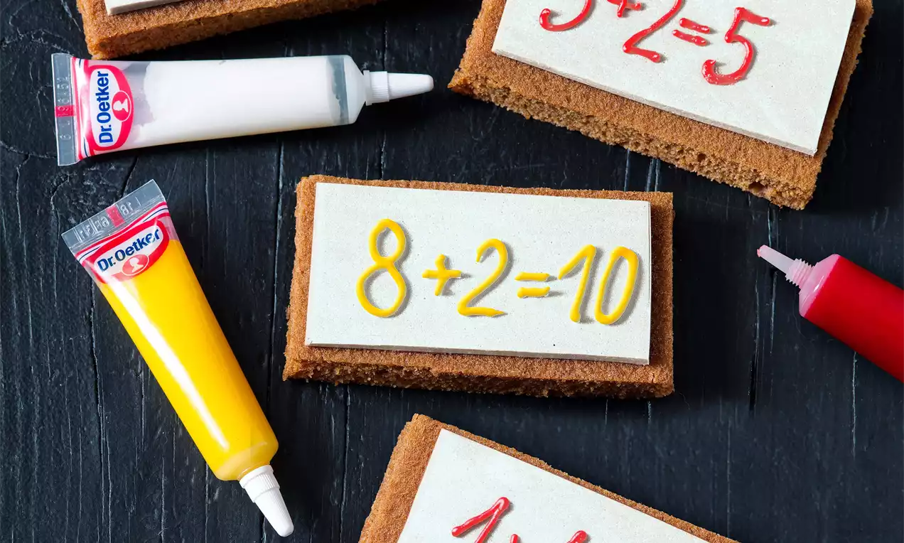 Hnědé sušenkové tabulky s bílou polevou, na které jsou barevnými tubami napsány matematické příklady, uspořádané na tmavém podkladu