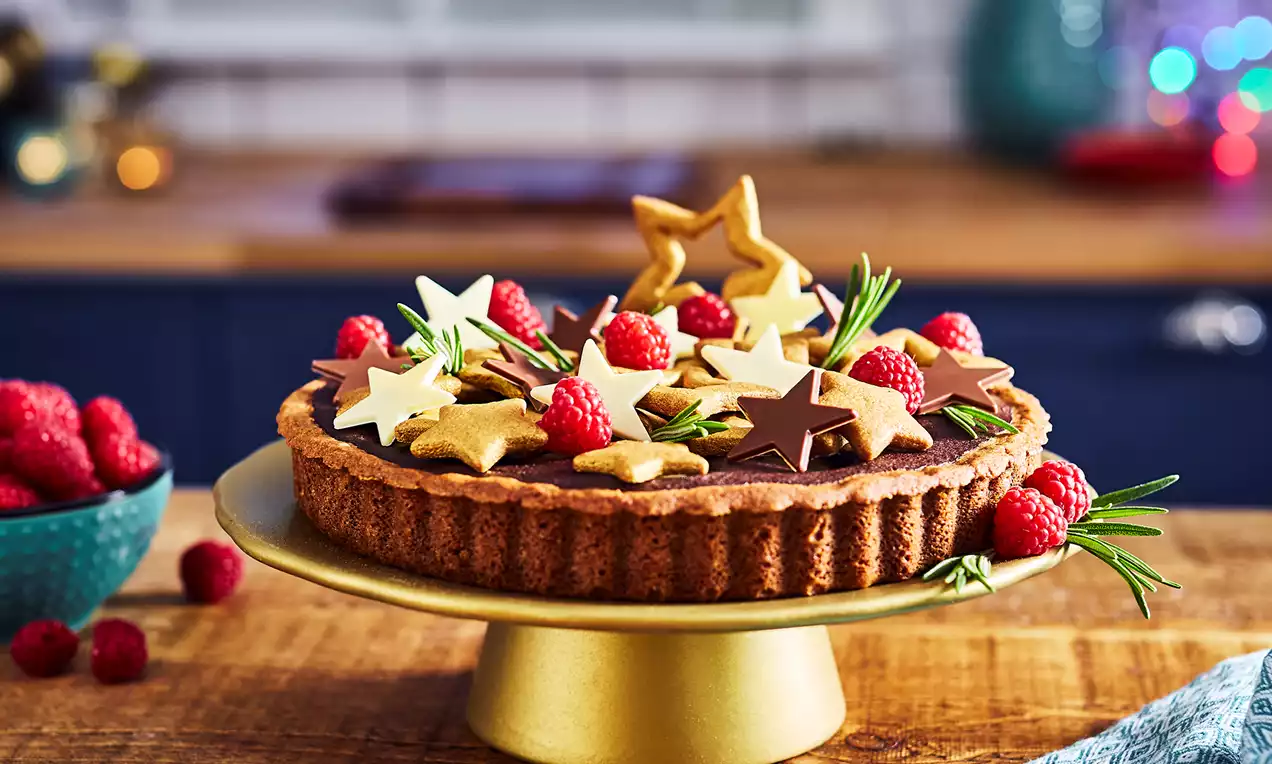 Golden-brown tart with scalloped edges topped with chocolate and white stars, sprigs of rosemary and bright red raspberries on a gold stand
