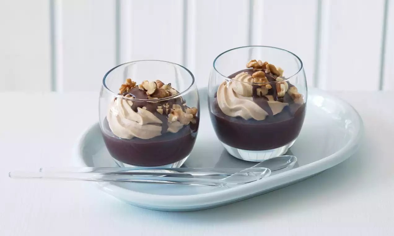 Dva sklenené poháre s vrstvami tmavohnedého krému, svetlého našľahaného piškótového krému, čokoládovou polevou a posypom orechov
