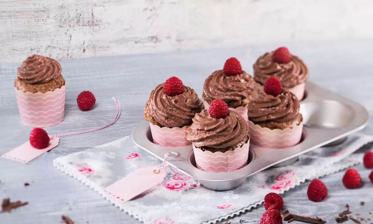 Vegane Schoko-Cupcakes mit cremigem, geschwungenem Topping, dekoriert mit tiefroten Himbeeren in rosa Förmchen auf einem Metallblech