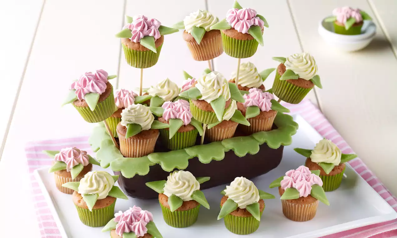 Zeleni papirnati omoti cupcakesa ukrašeni kremom boje bjelokosti i nježno ružičastom, raspoređeni na tamnosmeđoj podlozi okruženoj zelenim listićima