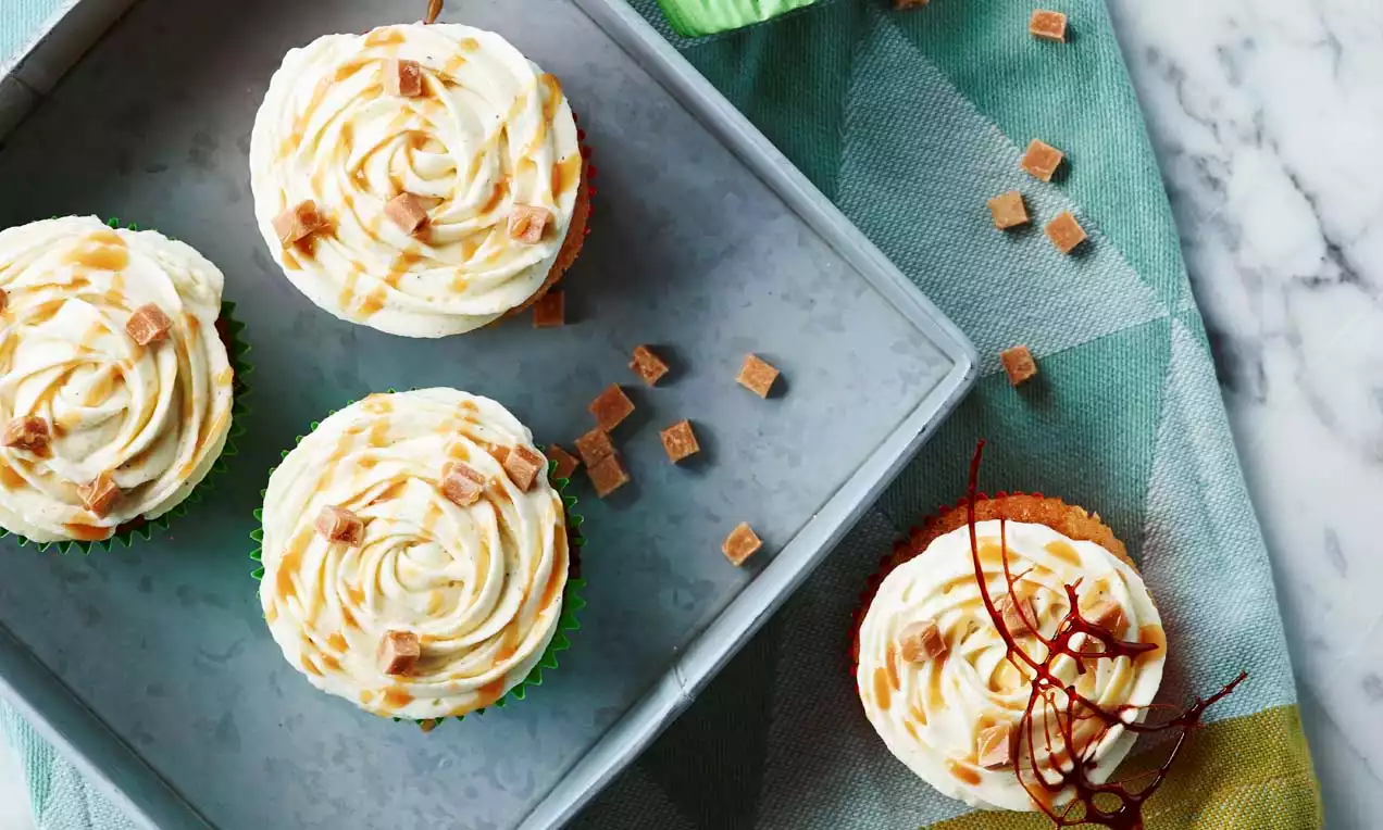 Vaaleat, pyöreät muffinssit metallisella, harmaalla alustalla, kermakuorrutuksen valkoiset spiraalit täplitettyinä kinuskiraidoilla ja toffeepaloilla