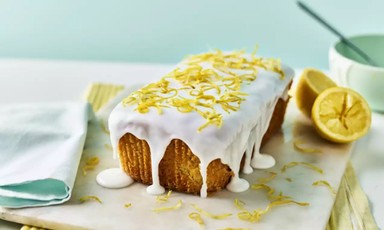 Golden rectangular loaf cake draped in glossy white icing with lemon zest scattered on top, set on a marble board with halved lemons