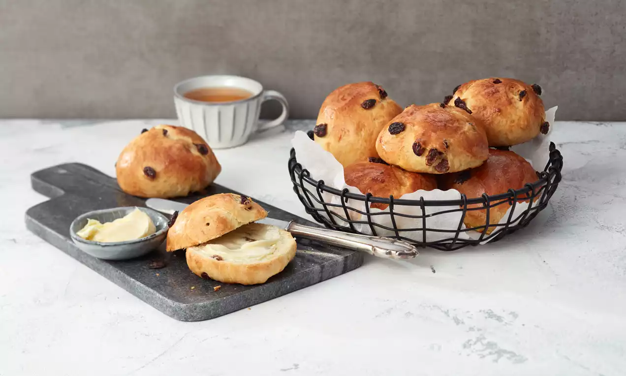 Runde, goldbraune Rosinenbrötchen mit glänzender Oberfläche in einem schwarzen Korb, dazu ein aufgeschnittenes Brötchen mit Buttercreme auf einem grauen Brett