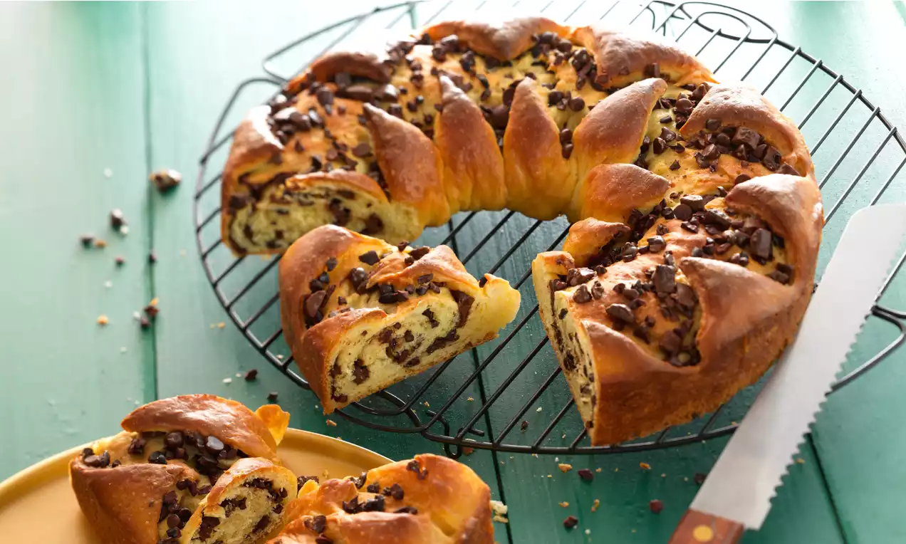 Aranybarna koszorúalakú kalács vékony szeletekre vágva, puha textúrával, csokoládédarabokkal hintve fémrácson zöld háttér előtt