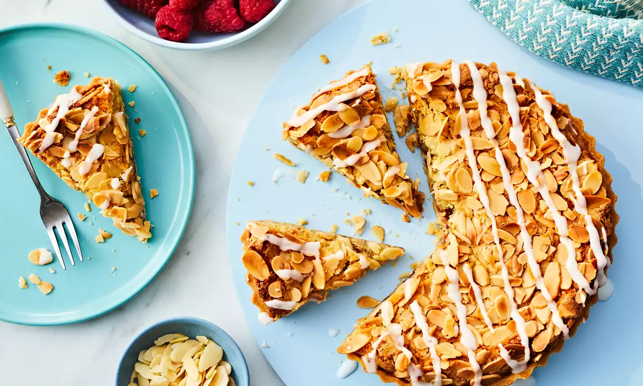 Bakewell Tart with golden pastry, almond flakes and white icing drizzles, sliced on a pale blue plate with raspberries nearby