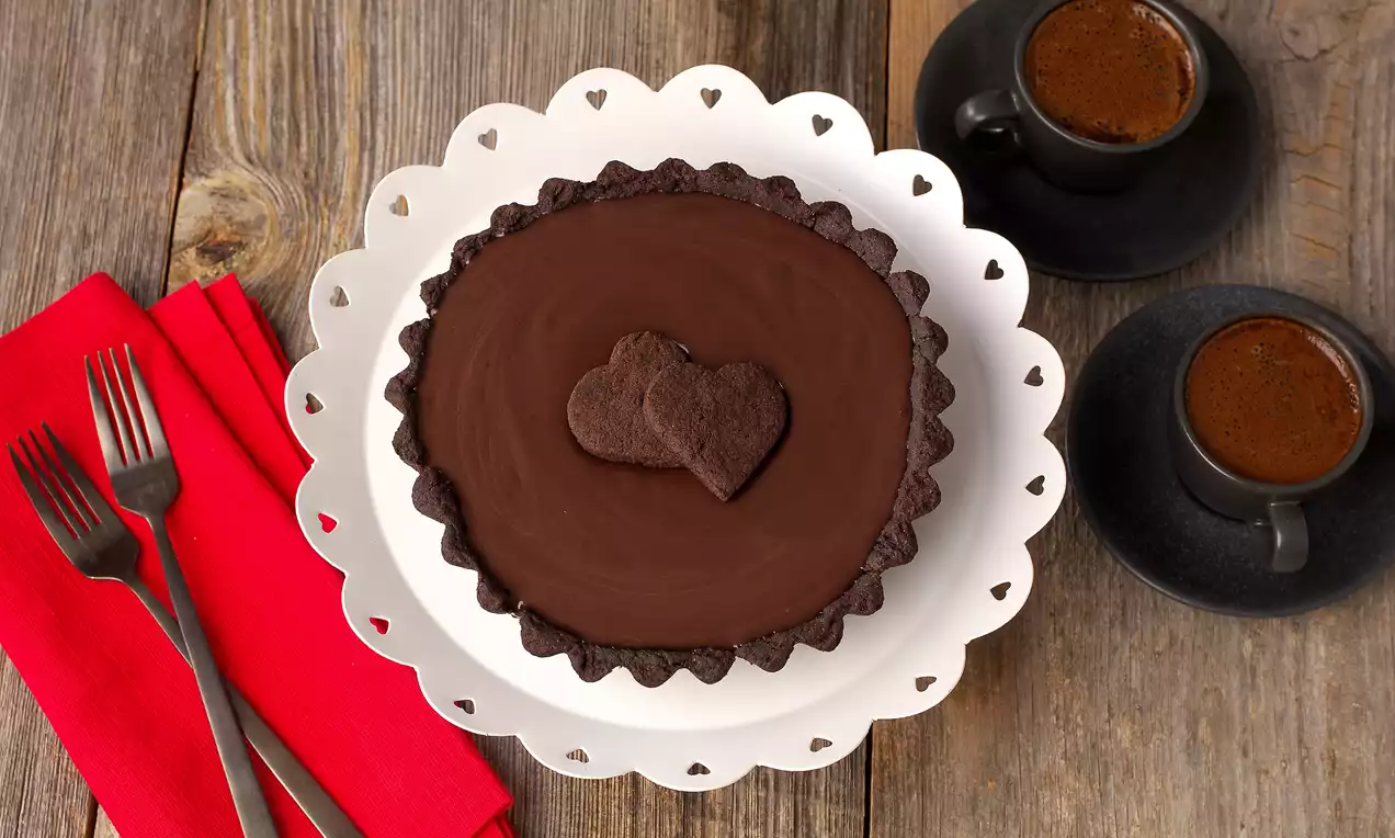 Ahşap bir zemin üzerinde beyaz kenar süslemeli tabakta sunulan, üstü pürüzsüz çikolata kaplı kalp şekli detaylı tart