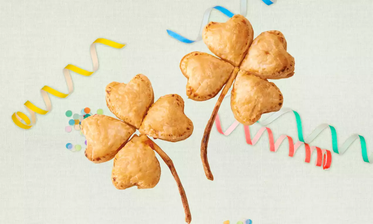 Goldbraune Kleeblätter aus Blätterteig mit zarten Herzblättern und dünnen Stielen, dekoriert mit bunten Papierstreifen auf hellem Hintergrund