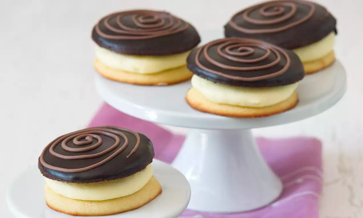 Kulaté dortíky skládající se ze světlého piškotu, krémové náplně a čokoládového povrchu zdobeného spirálou na bílém stojanu s růžovým ubrouskem