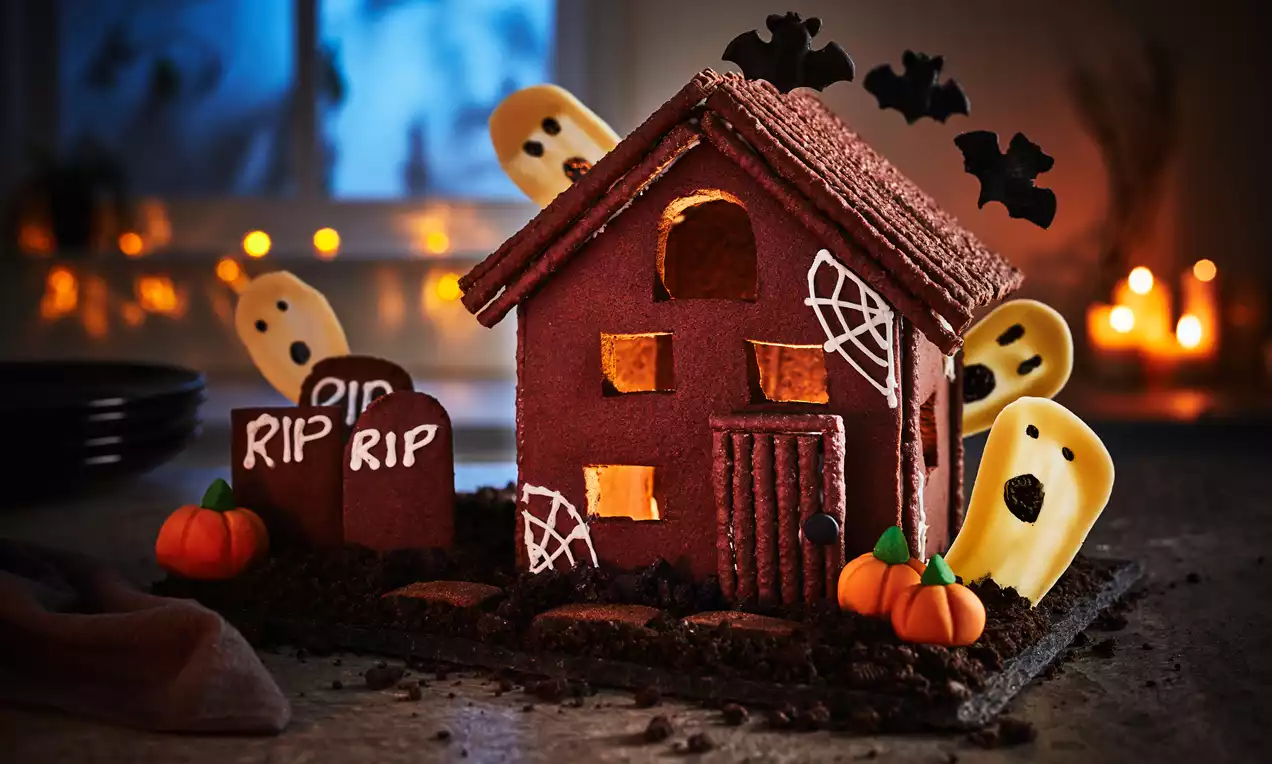 A dark chocolate biscuit house with textured walls, spooky windows, cookie gravestones, ghost shapes, pumpkins, and bat toppers on soil-like crumbs