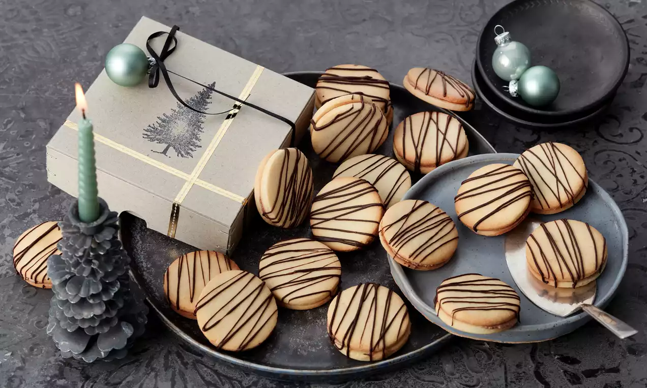 Runde Marzipan-Taler mit heller Oberfläche und dunkler Schokoladen-Dekoration auf grauen Tellern mit Kerze, Geschenkbox und Kugeln drapiert