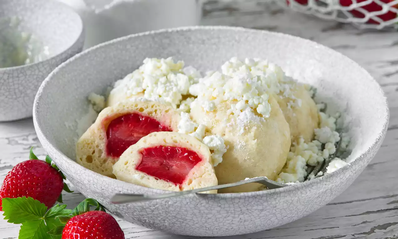 Světlé, kulaté kynuté knedlíky na texturovaném šedém talíři, jeden rozpůlený s červenou jahodovou náplní, posypané bílým tvarohem