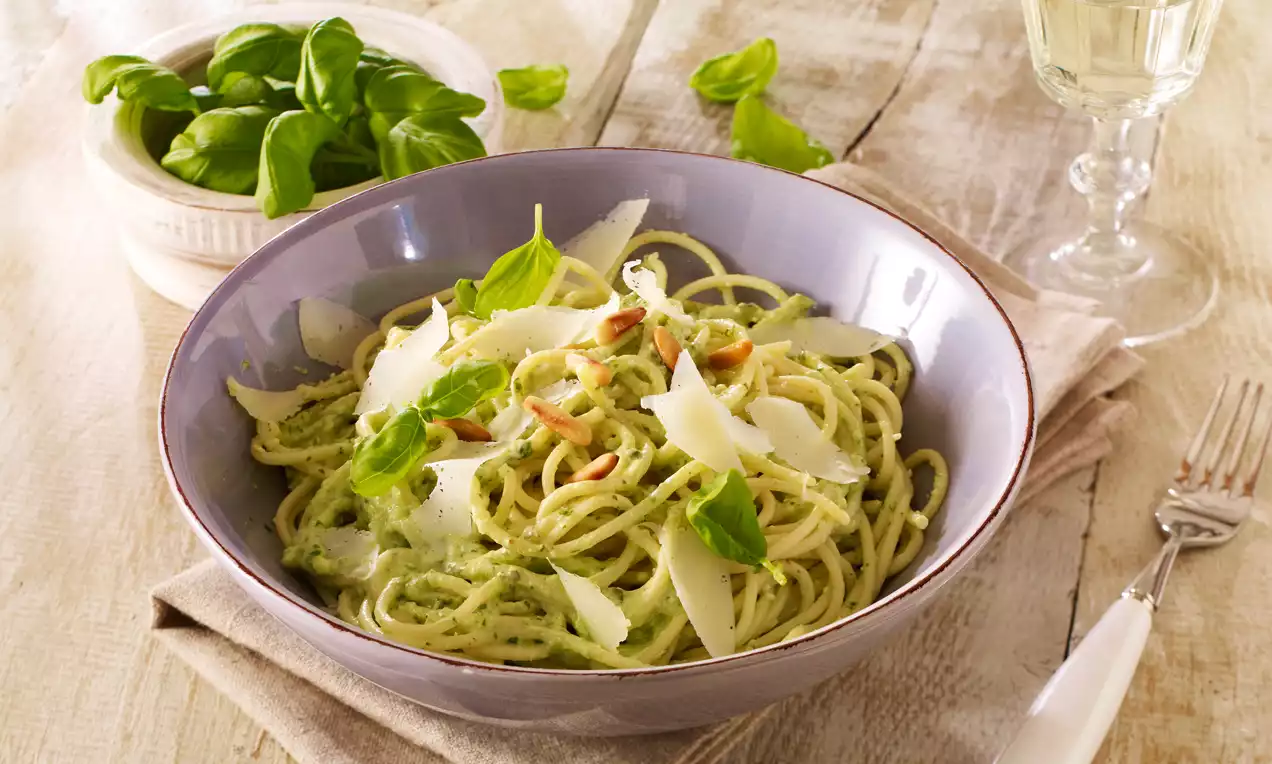 Spaghetti mit grüner Pestocreme, gerösteten Pinienkernen, frischen Basilikumblättern und Parmesanspänen in einer lila Schale auf Holzuntergrund