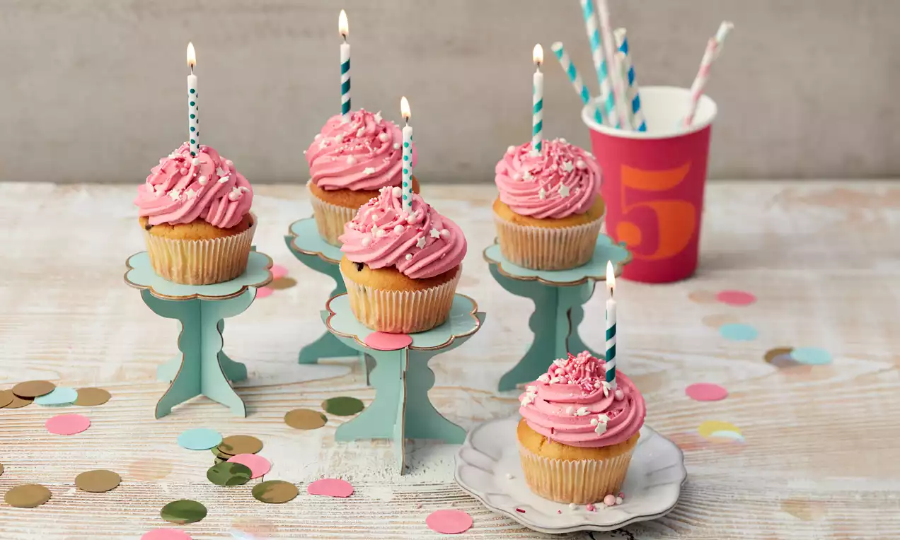 Geburtstags-Cupcakes mit rosa Cremetopping, weißen Streuseln und gestreiften Kerzen auf kleinen pastellfarbenen Ständern, dekoriert mit Konfetti