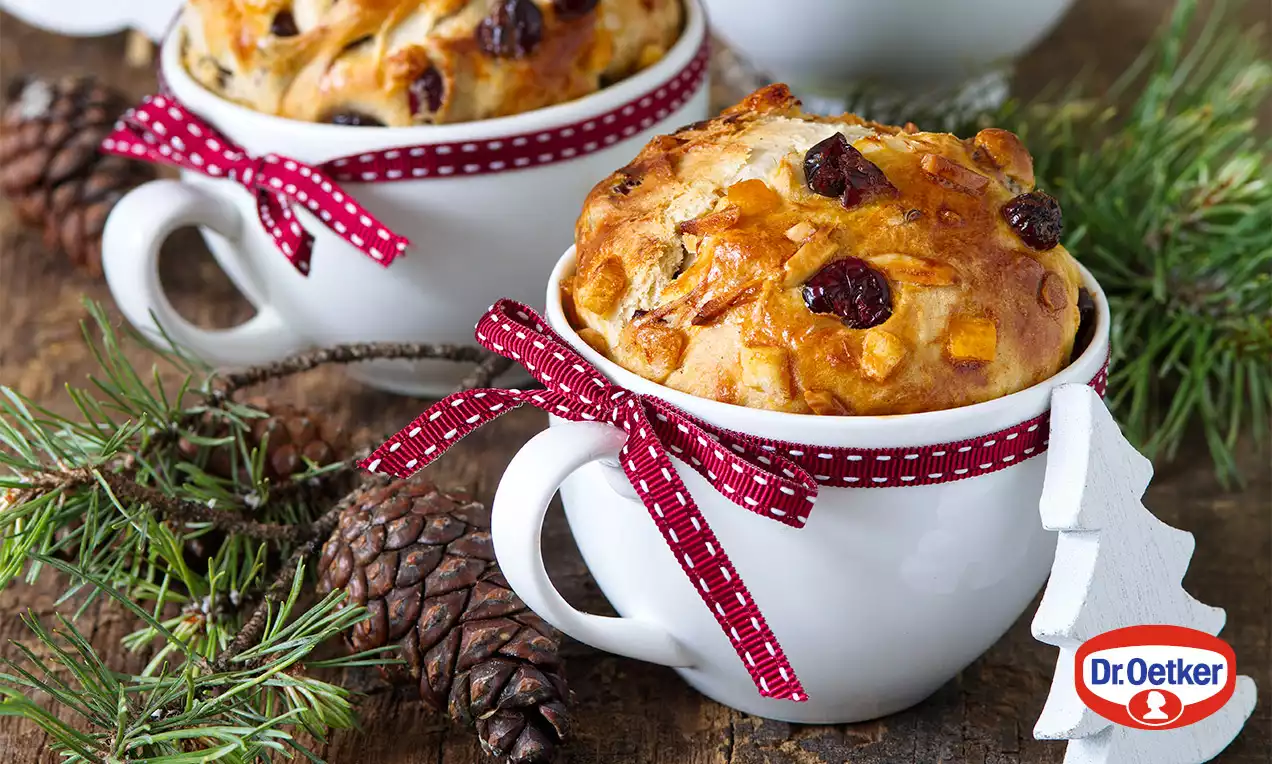 Zlatohnedé pečené buchtičky s brusnicami v bielych hrnčekoch ozdobených červenou stuhou, na drevenom stole s ihličím a šiškami