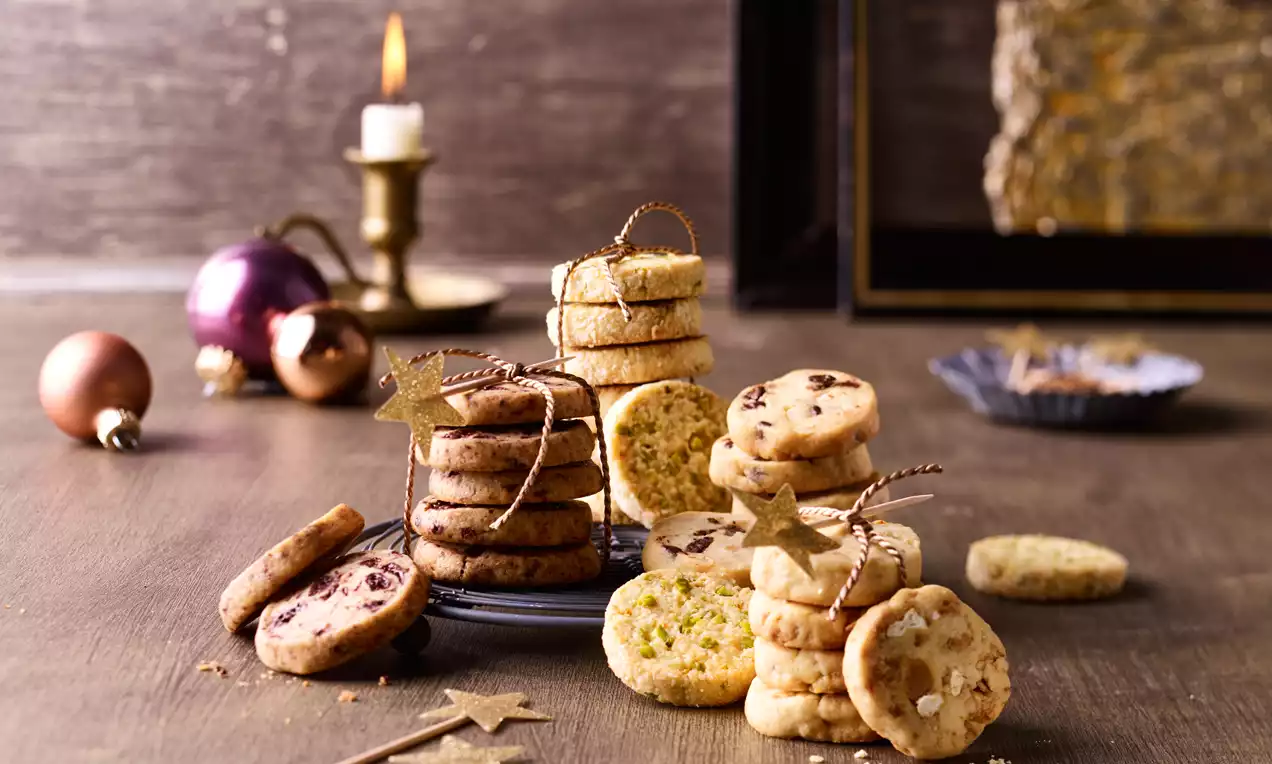 Mehrere Sorten rundes Gebäck in verschiedenen Farben und Texturen auf einem Holzuntergrund, teils gestapelt und mit goldenen Sternen dekoriert