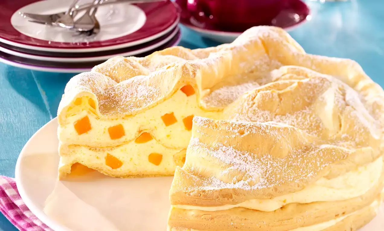 Brandteigtorte mit goldgelbem, fluffigem Teigdeckel, Aprikosenstückchen in hellgelber Cremefüllung, leicht mit Puderzucker bestäubt auf weißem Teller