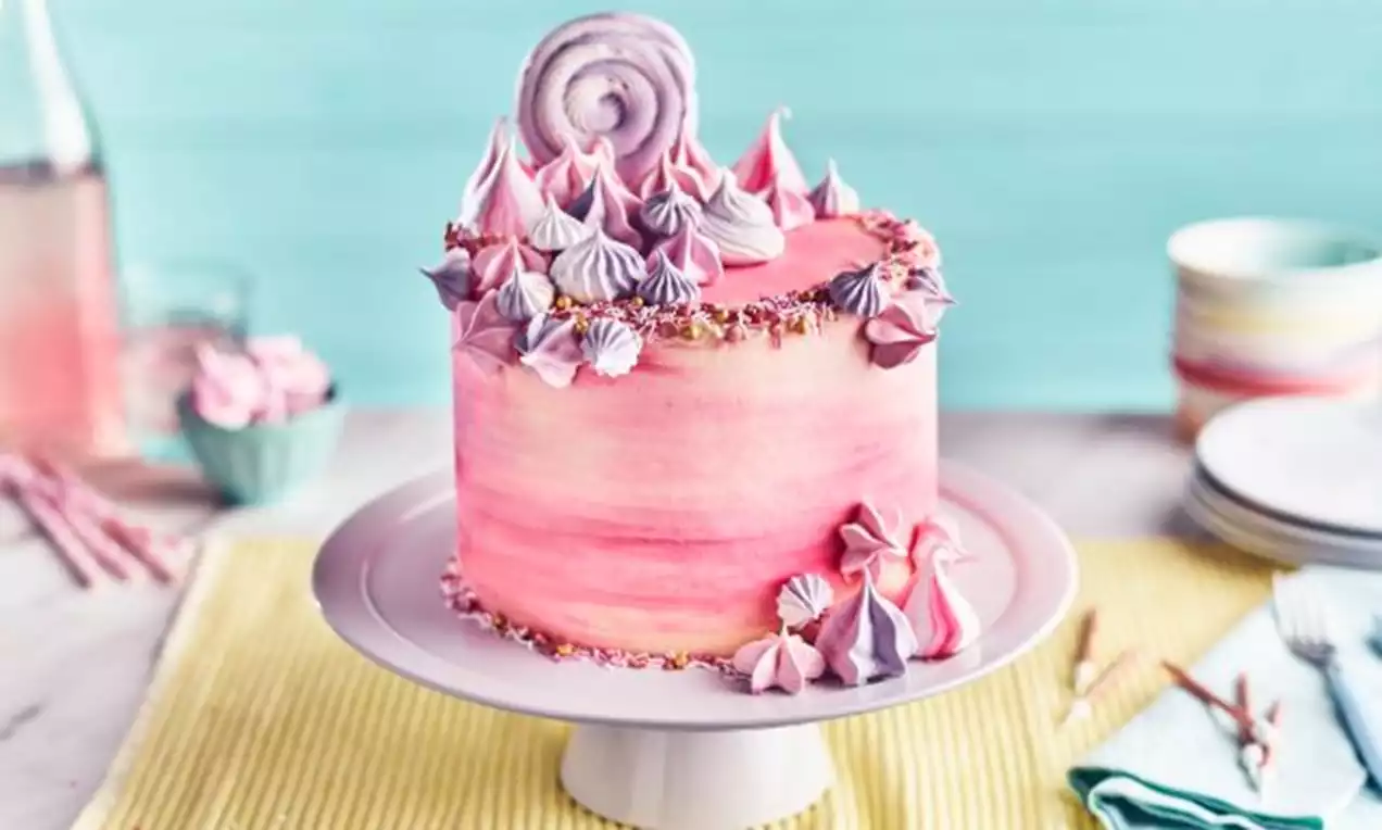 A tall layered pink and white meringue cake with swirled pastel peaks, set on a white stand against a bright pastel background