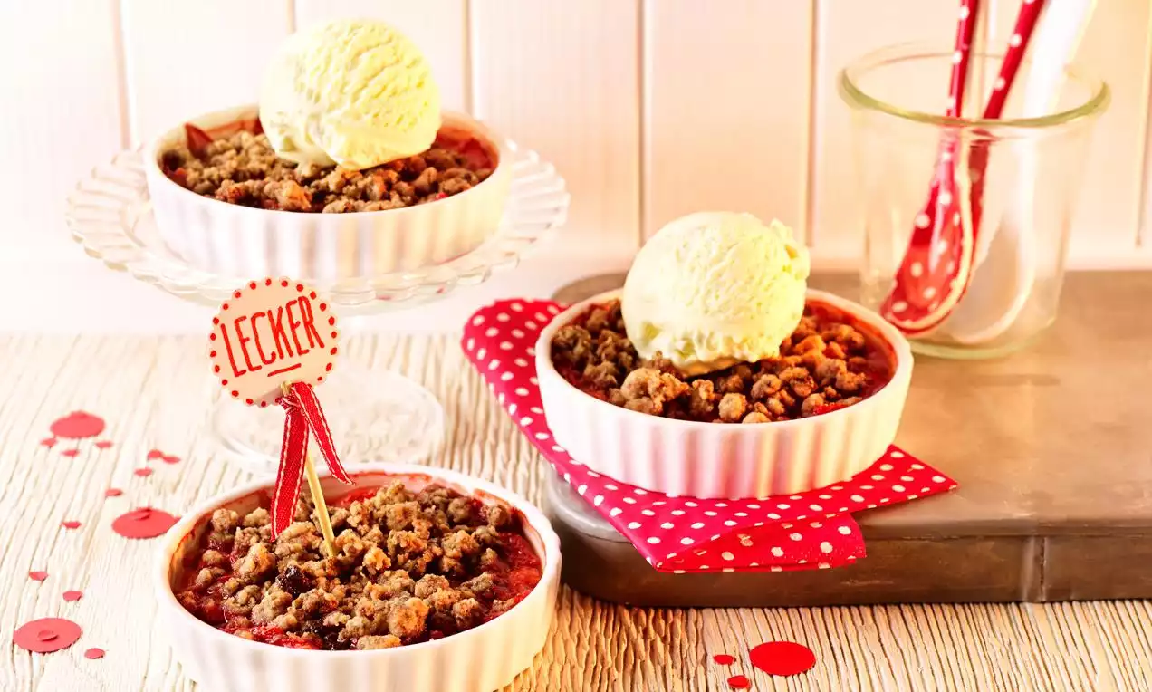 Crumble de căpșuni cu crustă aurie și căpșuni calde, servit în boluri albe, decorat cu înghețată vanilată pe fundal festiv cu accente roșii