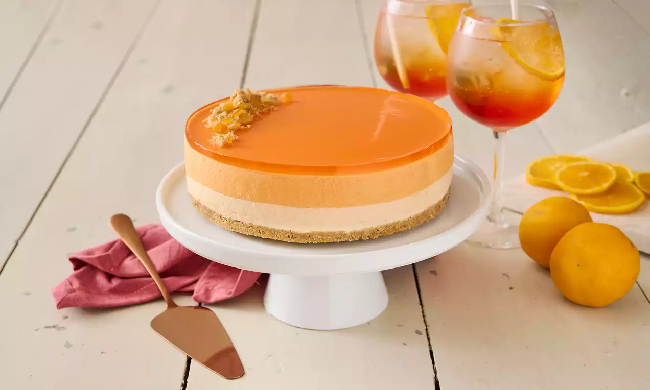 Aperol Spritz-cheesecake med orange gelatinöverdrag, ljusa krämiga lager, gyllenbrun kakbotten på vit bricka med citrus och cocktailglas