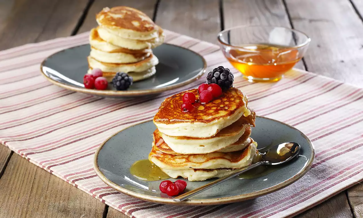 Ahşap bir zemin üzerinde serili çizgili bezin üstünde, gri tabaklarda istiflenmiş glutensiz pancake'ler, taze meyveler ve bal ile sunulmuş