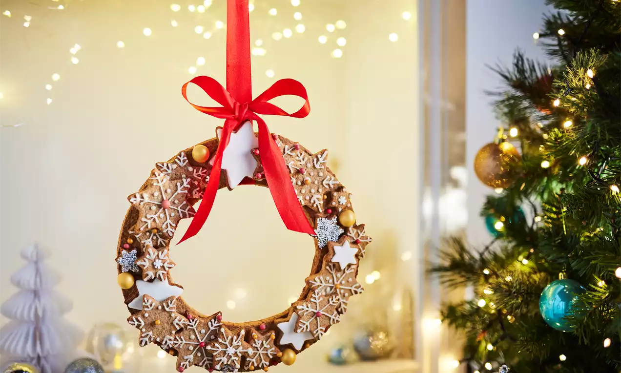 Golden gingerbread wreath studded with white iced snowflakes, gold and silver baubles, tied with a red ribbon, hanging by a Christmas tree