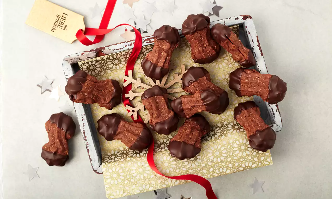 Nougatstangen aus brauner, geriffelter Masse mit dunkler Schokoladenglasur an den Enden, auf goldgemustertem Papier in einer weißen Holzschale