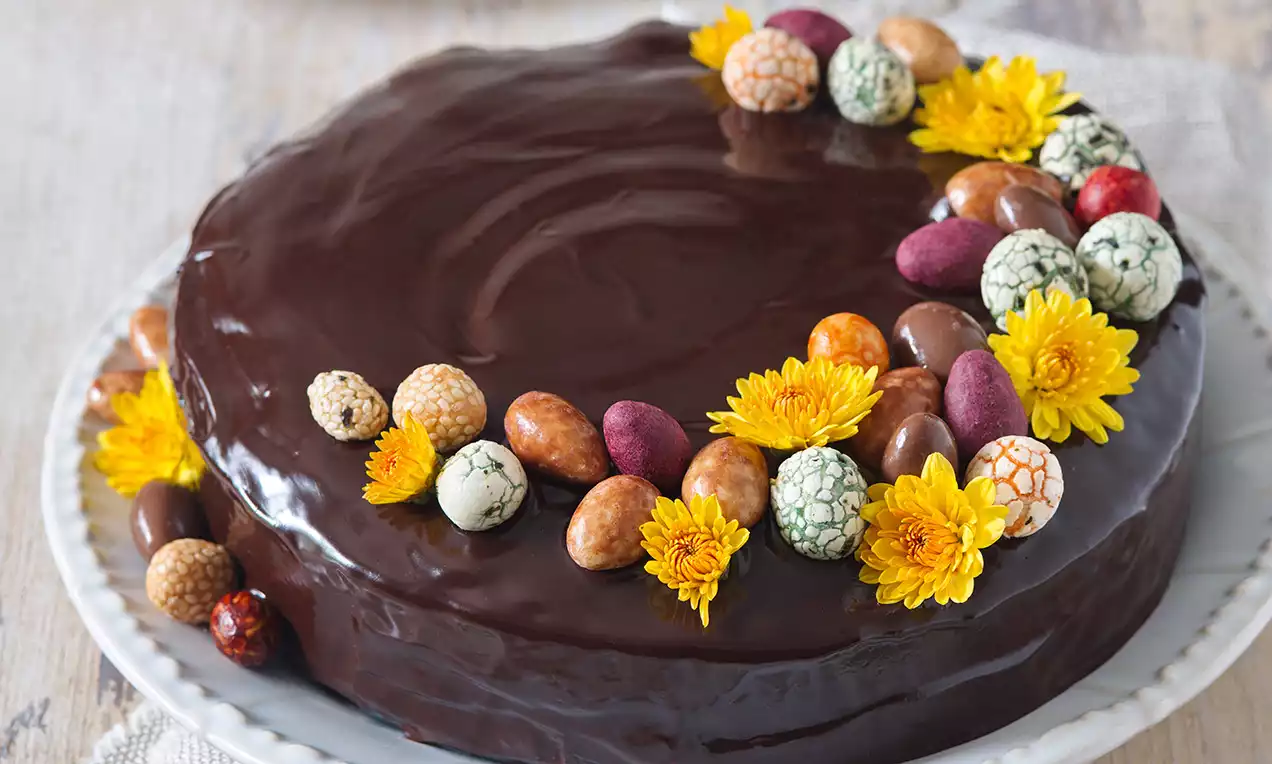 Tmavohnedá čokoládová torta s hladkým lesklým povrchom, ozdobená farebnými vajíčkami a žltými kvetmi na bielom tanieri