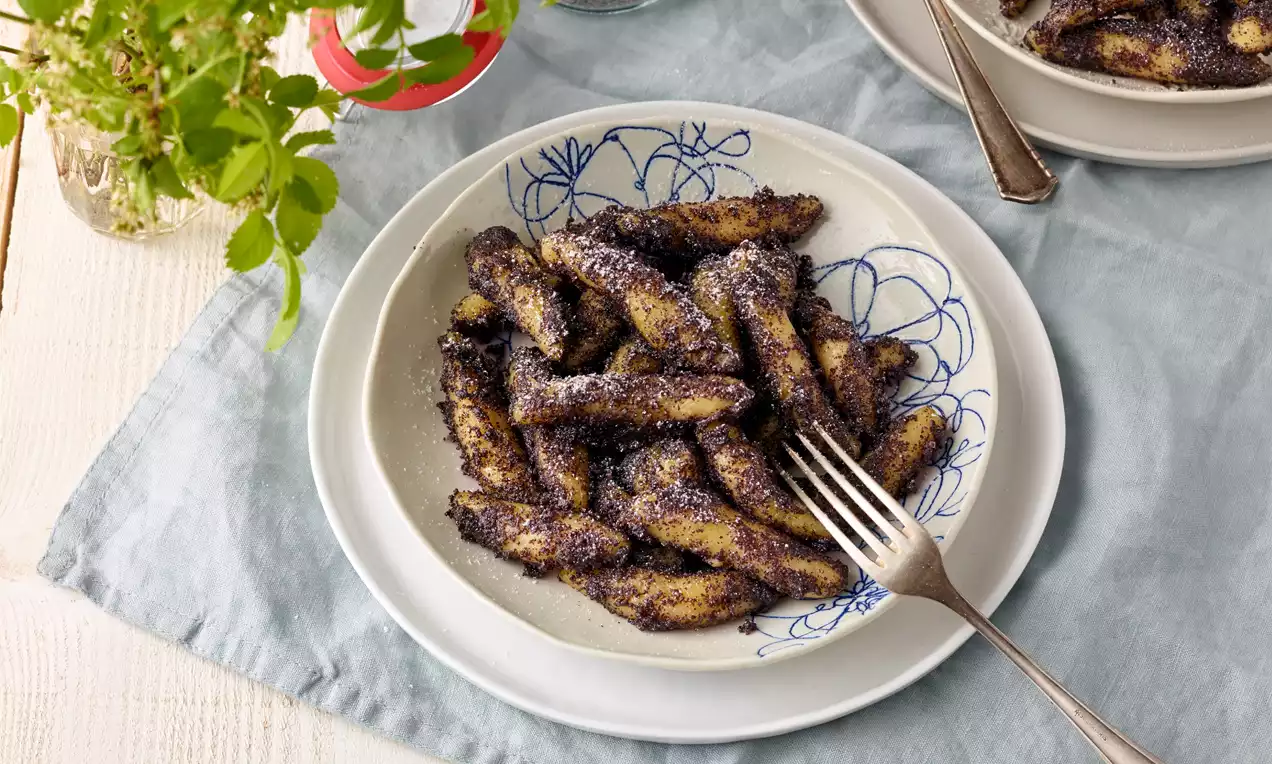 Mohnnudeln mit dunkler Mohnhülle und körniger Textur, locker angeordnet auf weißem Teller mit blauem Muster, daneben eine silberne Gabel