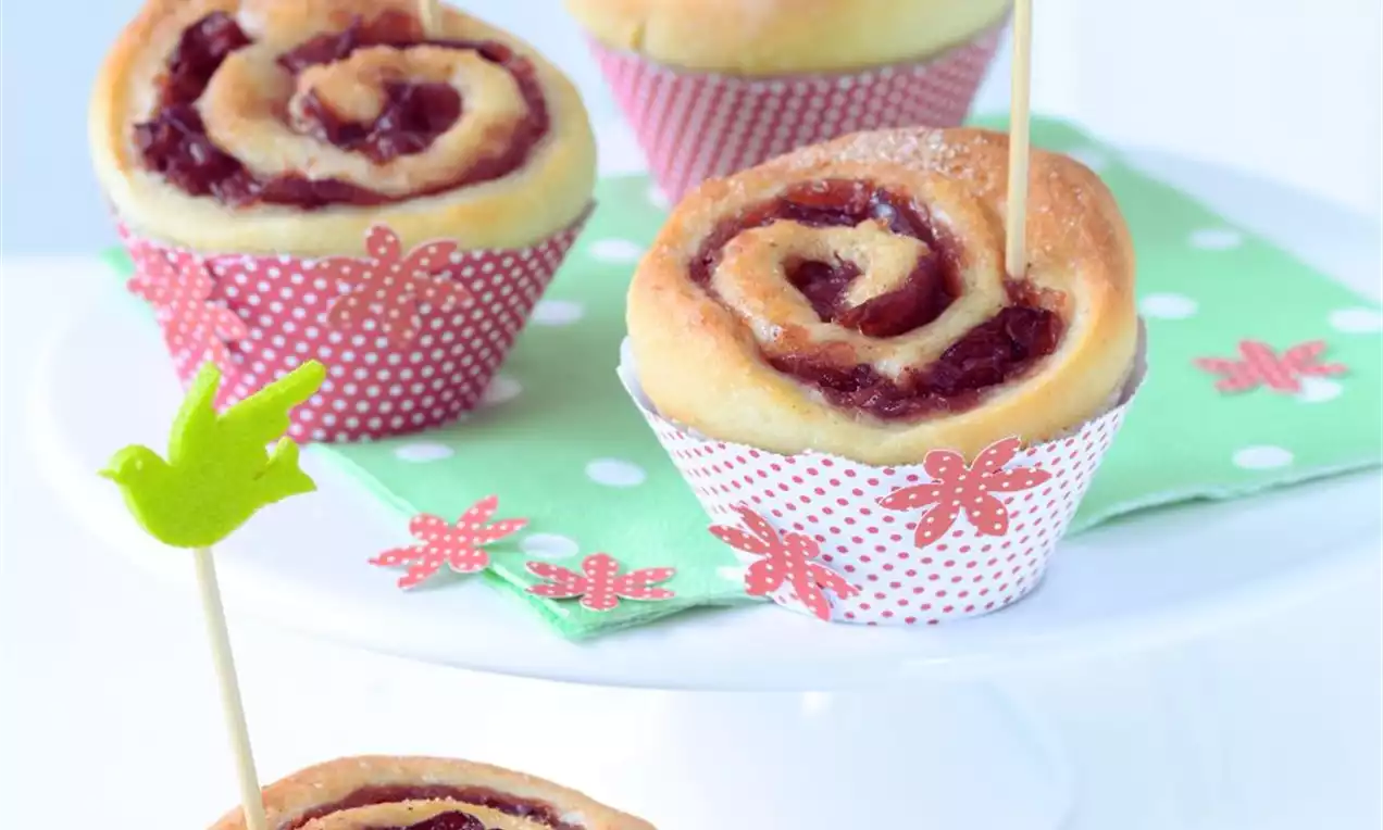 Světle hnědé kynuté pečivo stočené do tvaru růže s temně červenou náplní, podávané v puntíkovaných košíčcích s dekorativními barevnými ptáčky