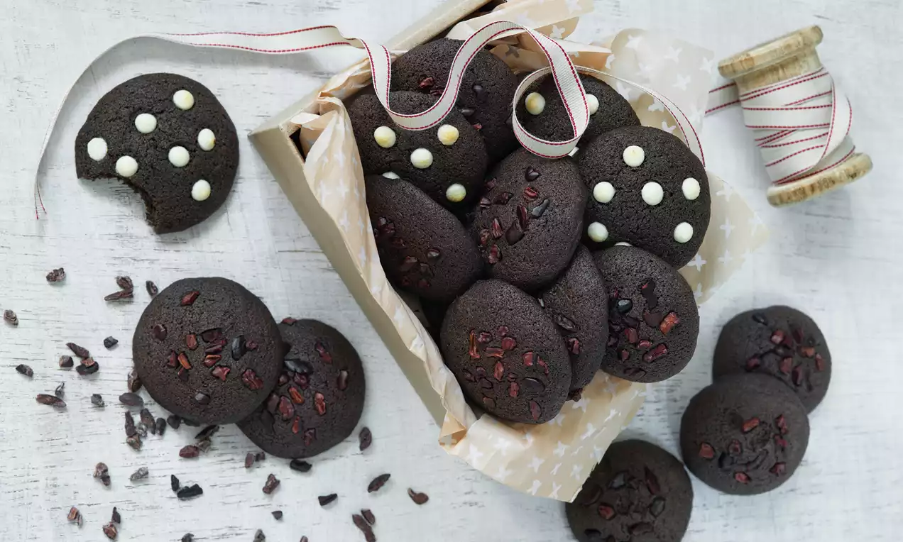 Tmavě hnědé kulaté cookies s nerovným povrchem posypané kousky kakaa, některé s bílými čokoládovými tečkami, na papíře v krabičce s mašlí