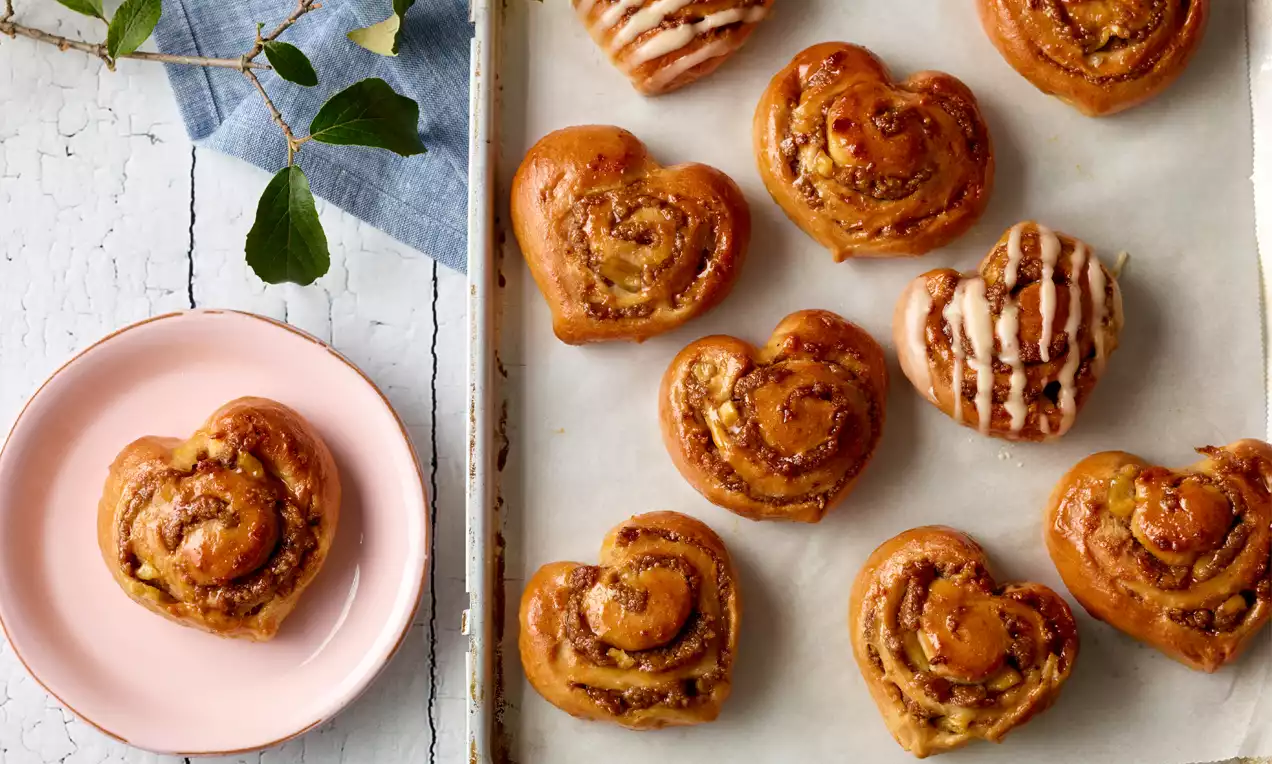 Herzförmige goldbraune Zimtschnecken mit glänzender Glasur auf Backpapier, dazu ein Teller mit einer Schnecke auf hellem Holzuntergrund