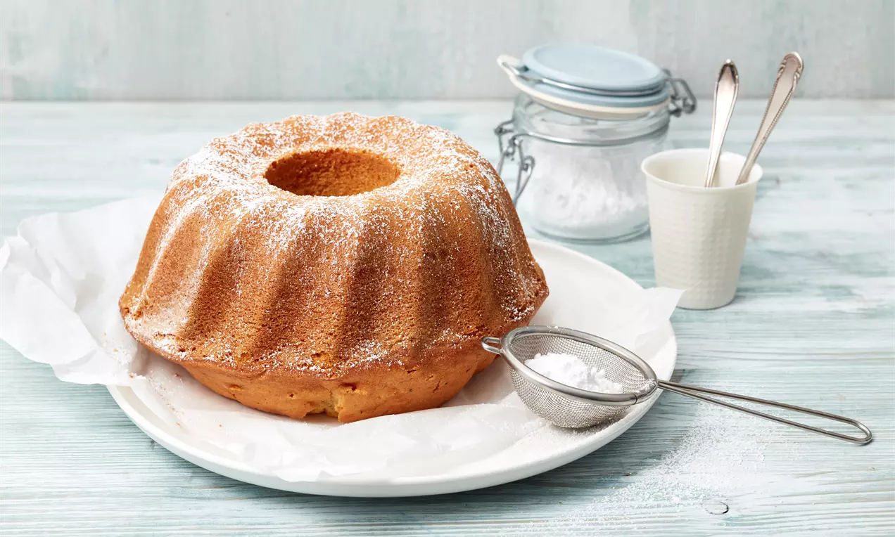 Rodonkuchen in Gugelhupfform mit goldbrauner Kruste, Puderzucker bestäubt und angeschnittene Stücke, die saftige, helle Krume zeigen