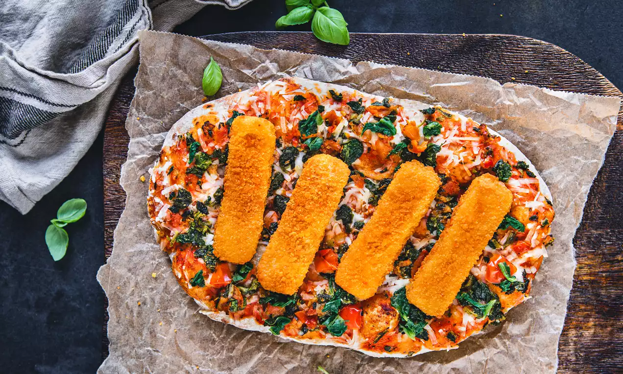 Rechteckige, goldbraune Fischstäbchen auf knuspriger veganer Pinsa mit roten Tomatenstücken, grünem Spinat und Kräutern auf braunem Backpapier