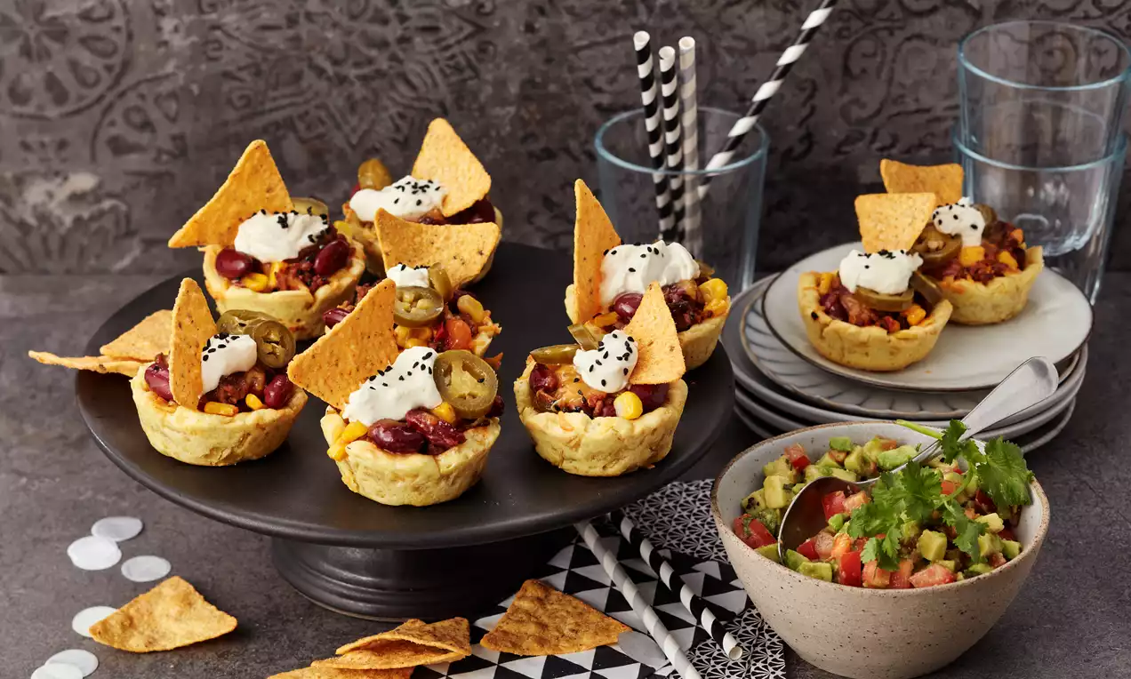 Goldbraune Taco-Muffins mit bunter Füllung aus Mais, Bohnen und Jalapeños, dekoriert mit Tortilla-Chips und Saucen-Tupfen auf dunklem Teller